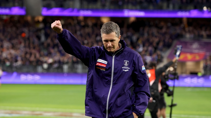 A man wearing a purple Fremantle Dockers jacket gives a clenched fist salute as he walks across an oval.