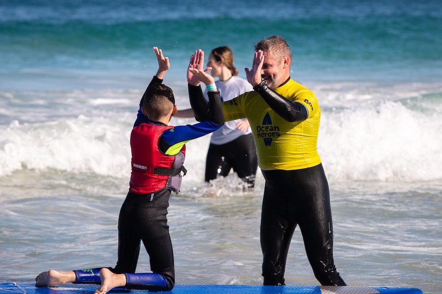 A young boy with a red life vest kneels on a surfboard high-fiving a man.