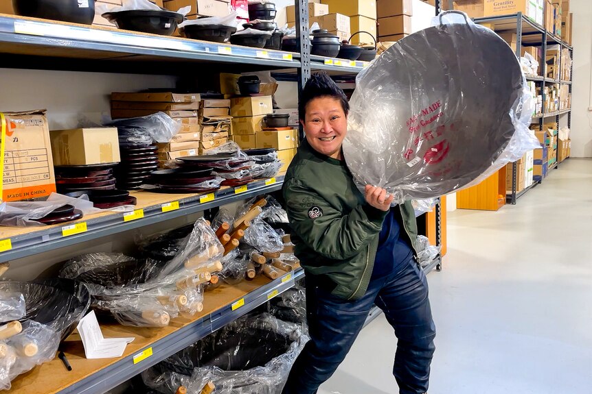 Chef Jerry Mai  holds up a large silver wok wrapped in plastic, next to a shelf full of them at the supermarket.