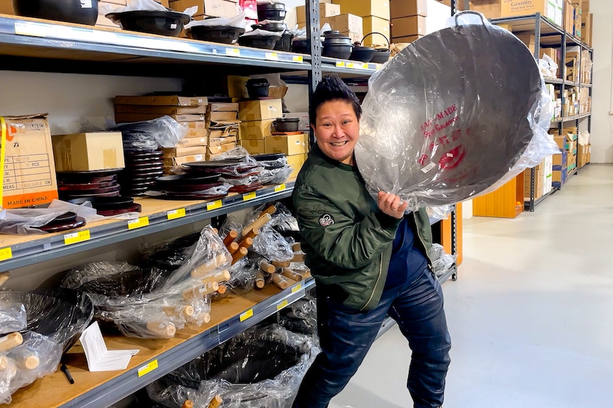 Chef Jerry Mai  holds up a large silver wok wrapped in plastic, next to a shelf full of them at the supermarket.