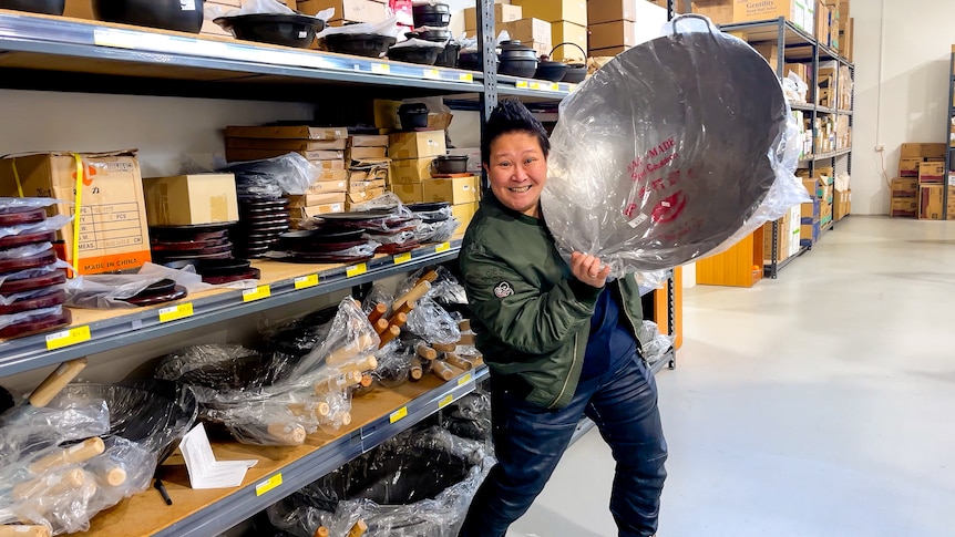 Chef Jerry Mai  holds up a large silver wok wrapped in plastic, next to a shelf full of them at the supermarket.