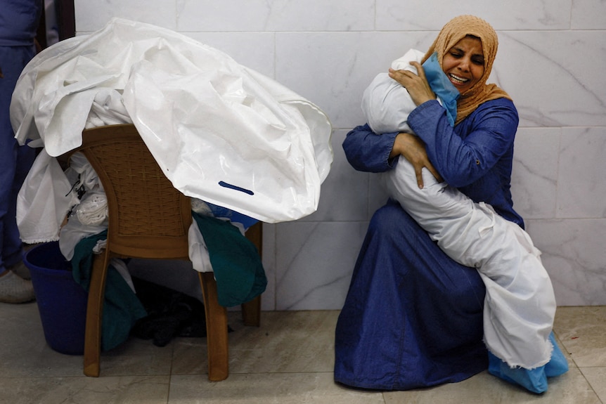 An anguished woman crouching in a hallway clutches a small body wrapped in a sheet to her chest.