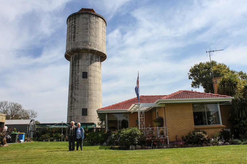 Two elderly people stand outside their home with an Australian flag and a water tank.