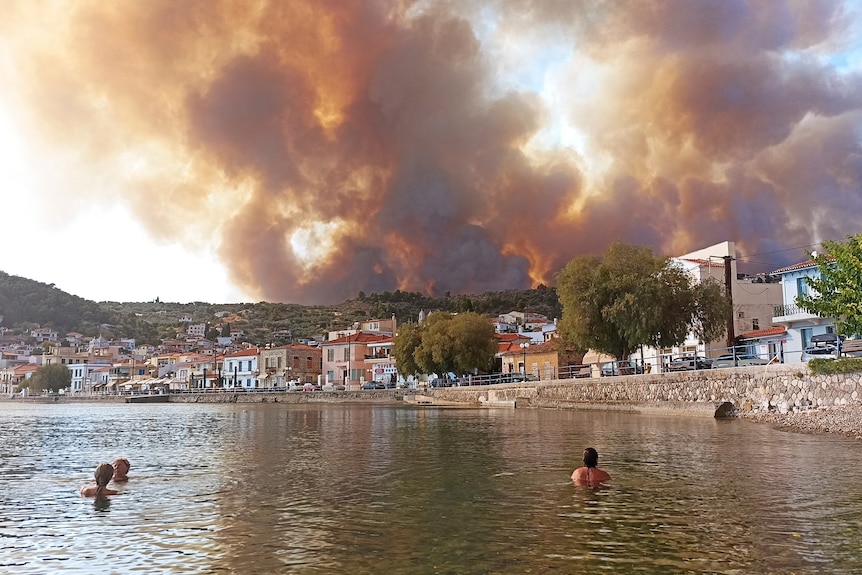 The heads of people swimming bob above the water looking at deep red clouds over a small greek town
