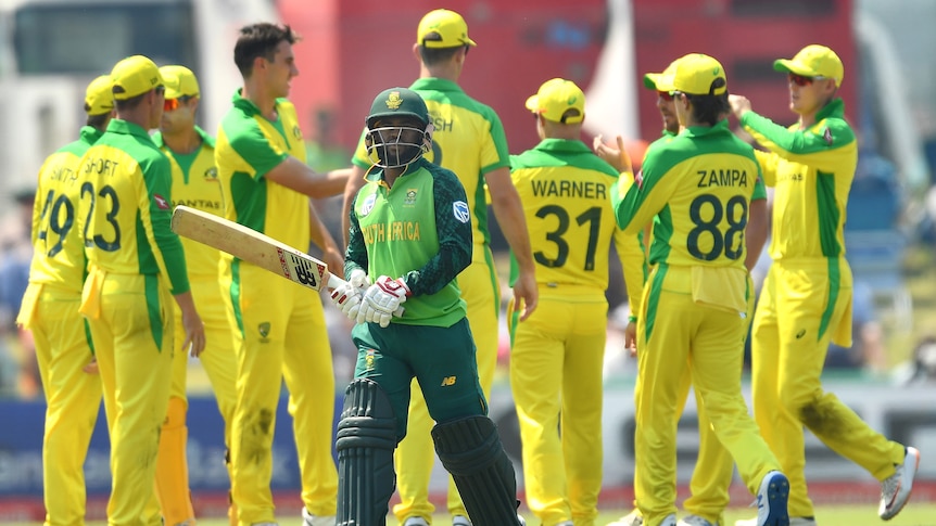 A South African batsman walks off holding his bat in both hands as the Australian ODI team celebrates in the background.