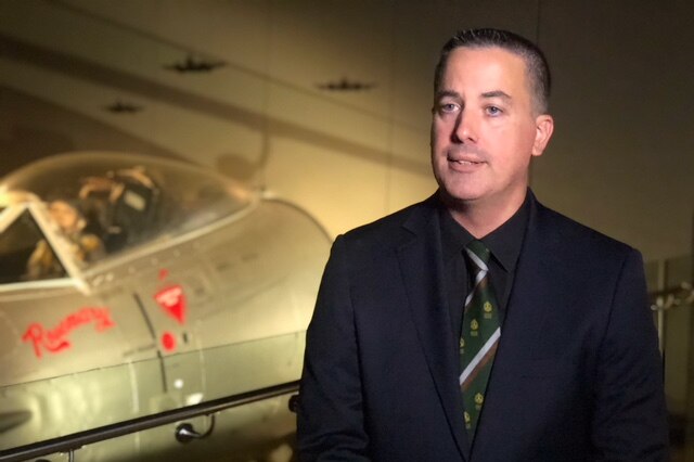 Michael Kelly wears a navy suit with a green, brown and white tie. He is mid-sentence. An old plane is in the background.