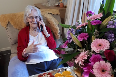 An elderly woman sits with a phone to her ear, surrounded by bunches of flowers and a platter of fruit. 