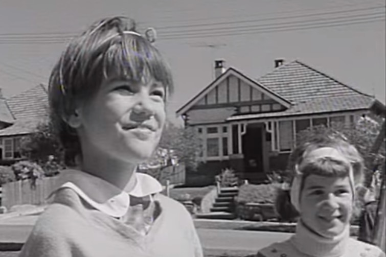 Still from black and white footage. A young girl with short hair smiles on a suburban street, with a friend in pig-tails