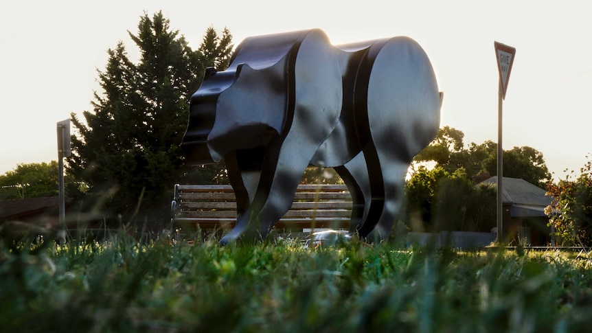 A large, black cat sculpture in with trees and a fading sunset behind, and grass in the foreground.