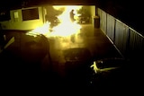 The silhouette of a hooded man in flames running through a carport.