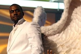Kanye West dressed in a white tuxedo with large angel wings. The crowd below him is raising their hands up to him.