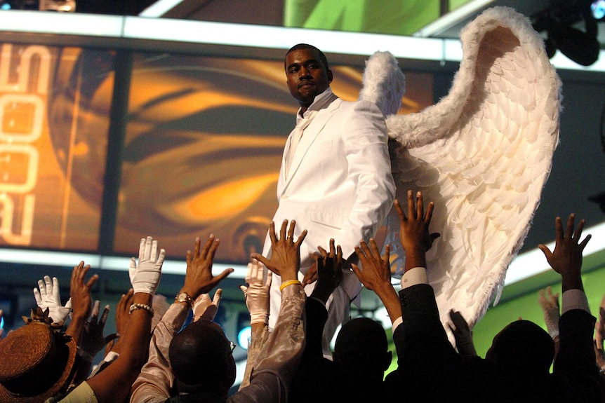 Kanye West dressed in a white tuxedo with large angel wings. The crowd below him is raising their hands up to him.