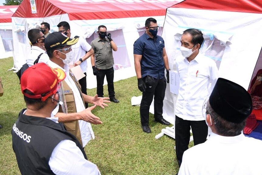 Indonesian President Joko Widodo wearing a mask listens as a relief official talks and gestures in front of relief tents.