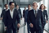 Characters of HBO show Succession walk down a hallway in dark business suits, eyeing each other