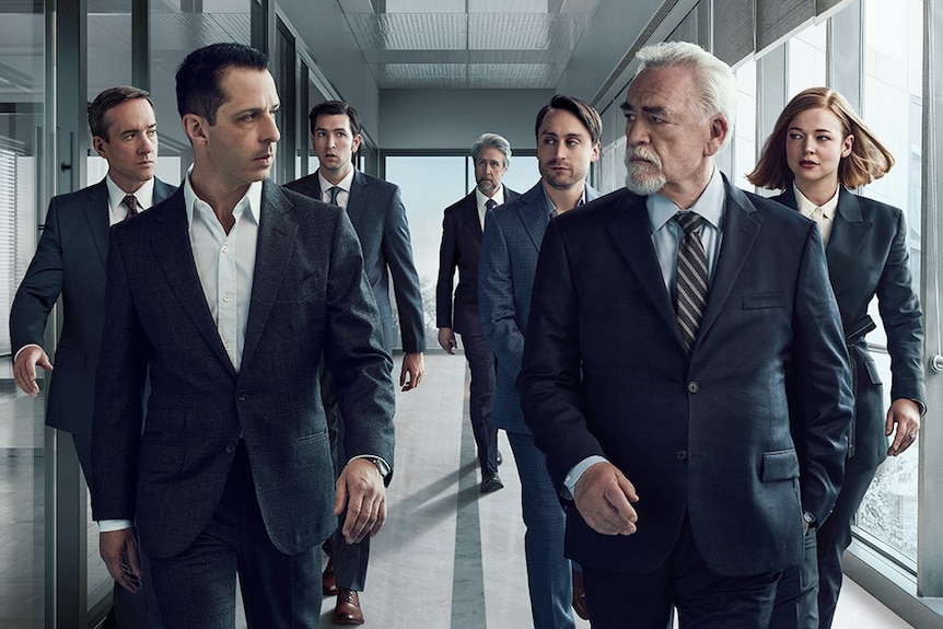 Characters of HBO show Succession walk down a hallway in dark business suits, eyeing each other