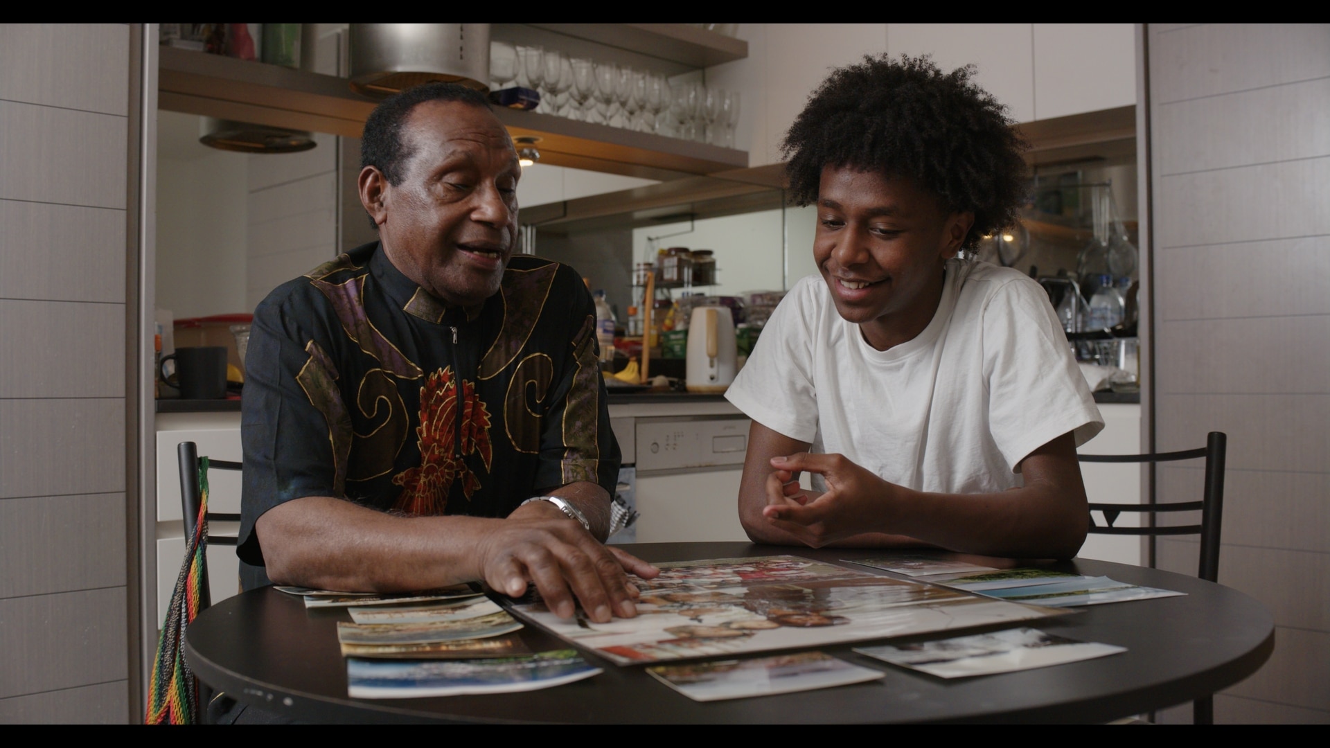 Two men seated at a small table and point to a picture board.