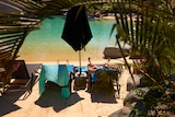 Two people lounge on deck chairs next to a tropical looking pool, a closed beach umbrella between them