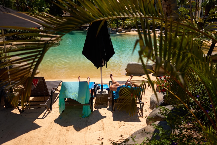 Two people lounge on deck chairs next to a tropical looking pool, a closed beach umbrella between them