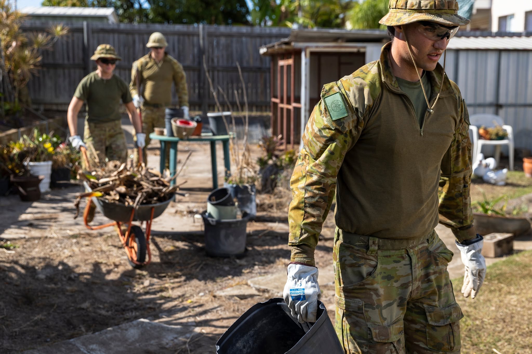 Soldiers in uniform carrying buckets and full wheelbarrows out of a backyard.