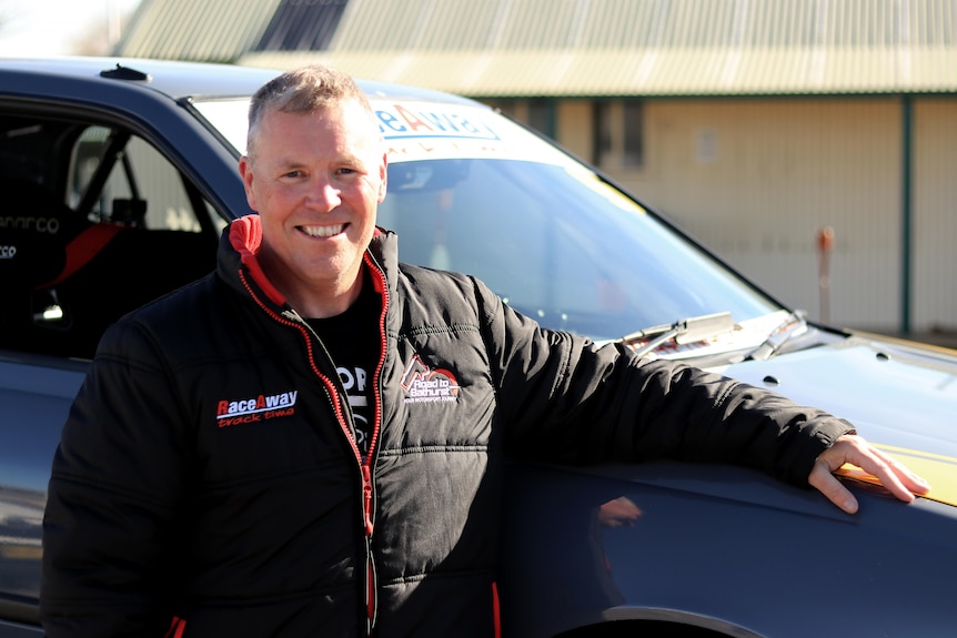 A smiling man with grey hair stands with a red and black jacket on, resting his arm on a small racing car.