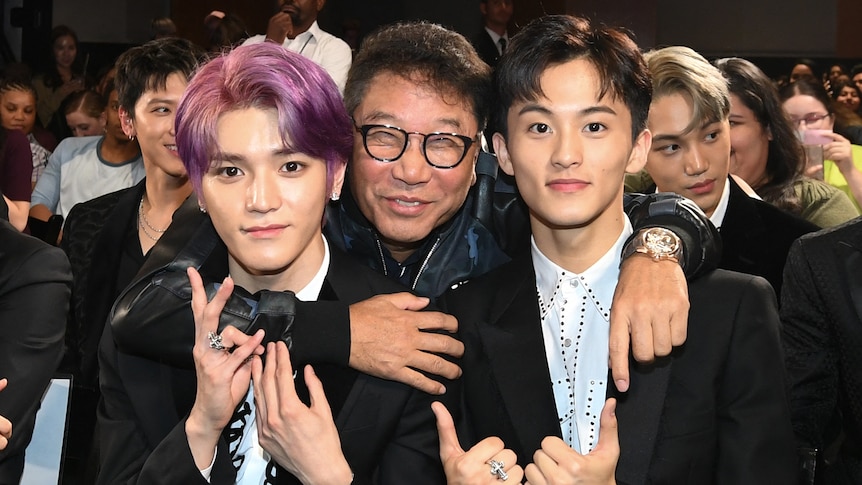 A middle-aged man in glasses smiles with his arms around two young K-pop stars, one with pink hair