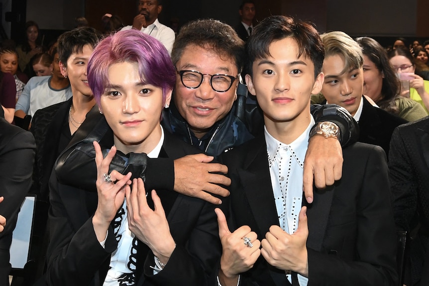 A middle-aged man in glasses smiles with his arms around two young K-pop stars, one with pink hair