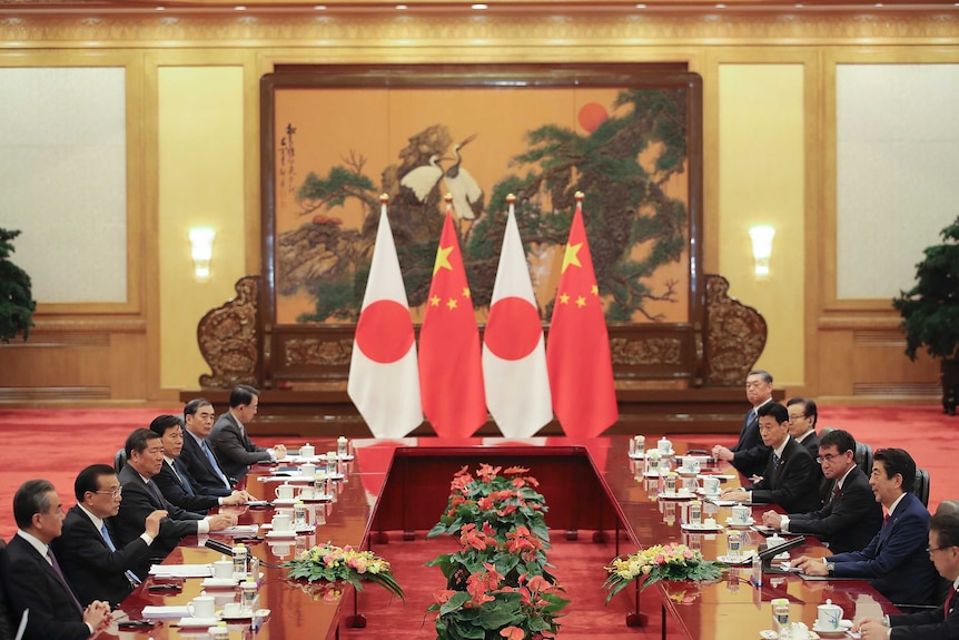 Chinese and Japanese government officials sit at large table with flags of their countries in the background