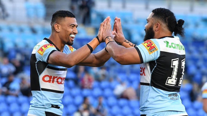 Two Polynesian Rugby players in light blue jerseys do a high ten salute with half empty seating area behind them.