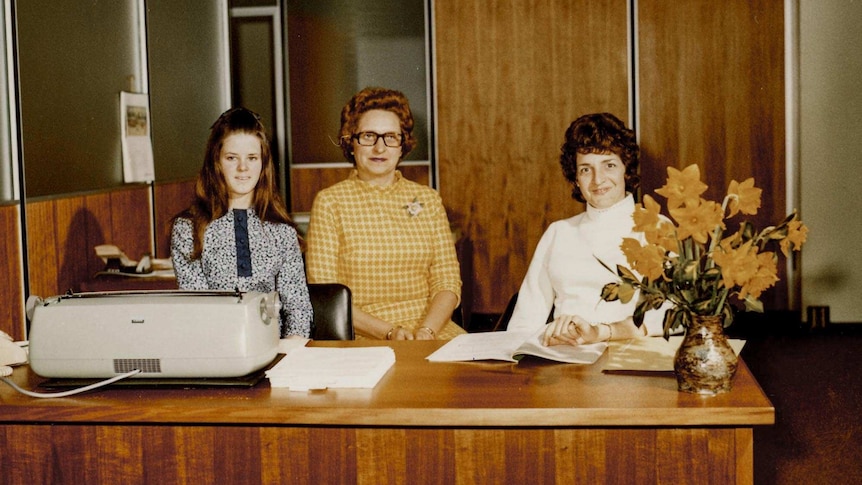 Three women sit in a wood panelled office behind a desk holding an electric typewriter