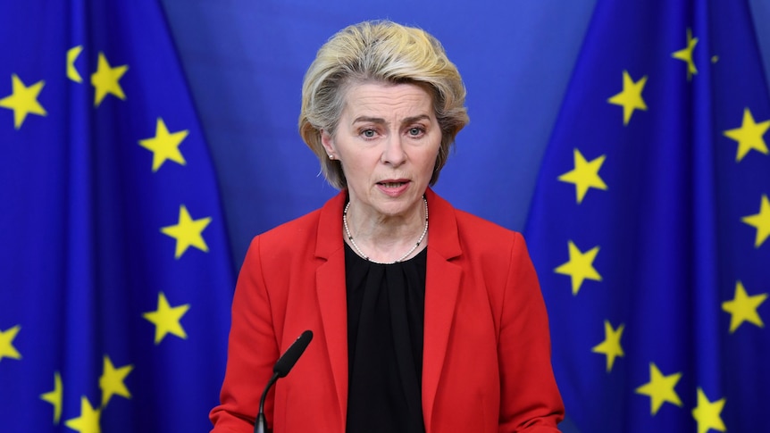 Ursula von der Leyen, an older blonde woman wearing a red jacket, stands at microphone in front of blue and yellow EU flag.