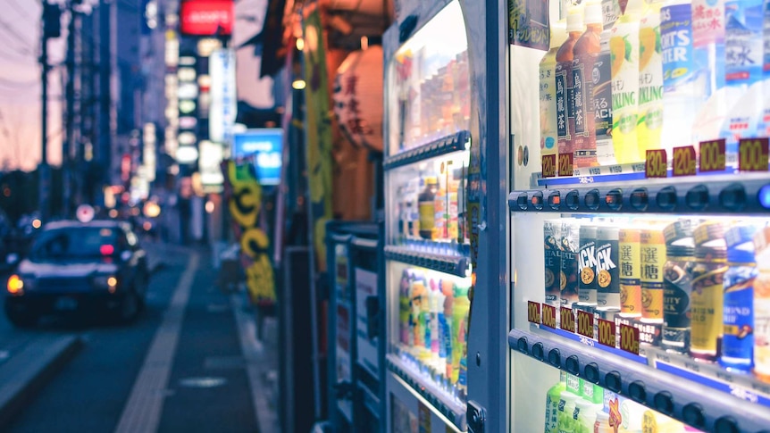 A Japanese street scene at evening with two neon-lit, brightly coloured vending machines, selling drinks, in the foreground.