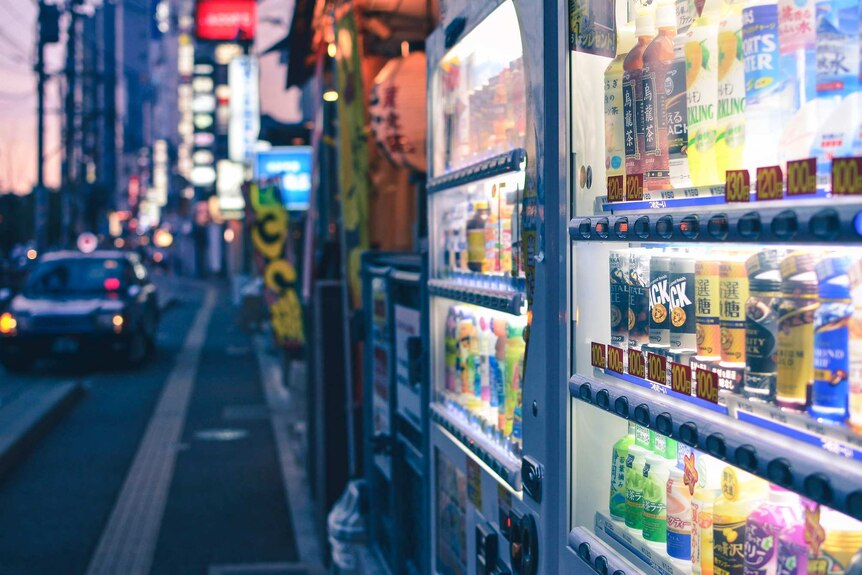 A Japanese street scene at evening with two neon-lit, brightly coloured vending machines, selling drinks, in the foreground.