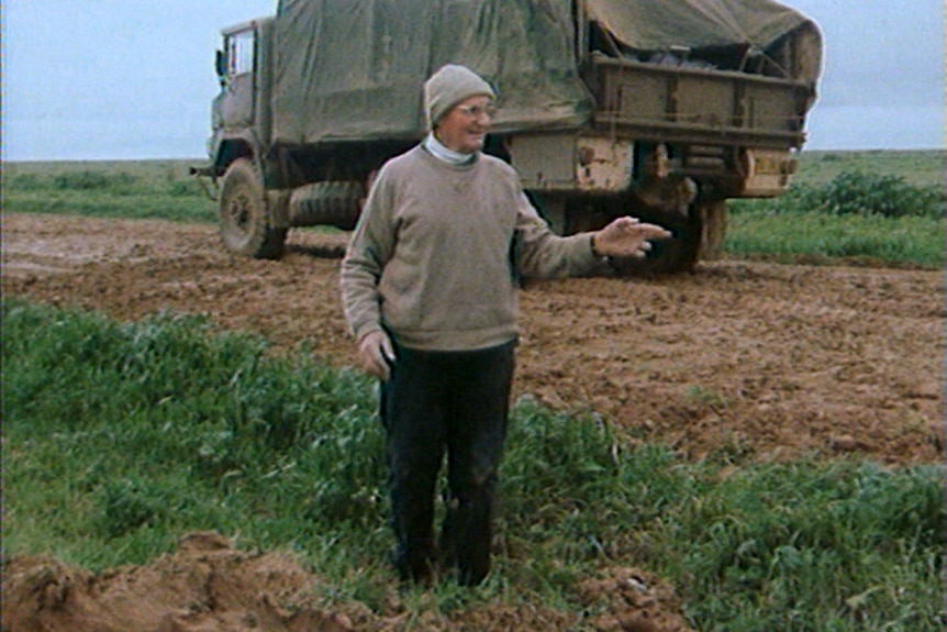 Bill Baird stands in front of his truck on a muddy stretch of road, he points towards the road behind the truck