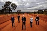 A group of five men stand in an outback setting. Their is red dirt all around them.