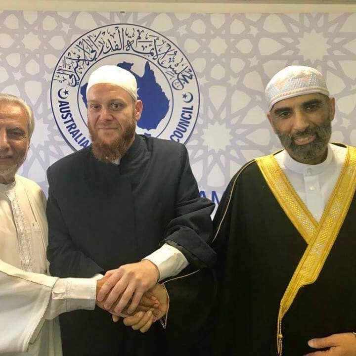 Three men in religious dress shaking hands in front of an Australian Islamic Council poster