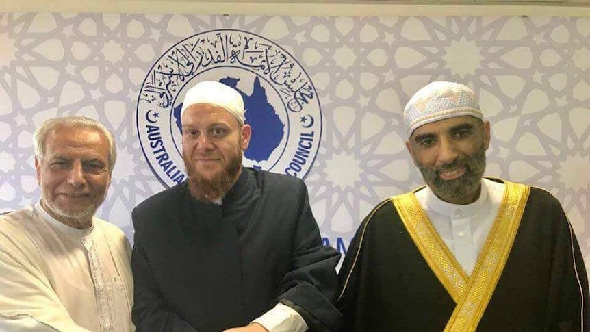 Three men in religious dress shaking hands in front of an Australian Islamic Council poster