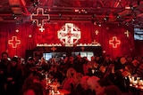 People eat at long tables under red lighting and decorative crucifixes.