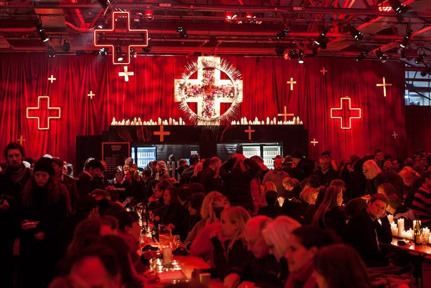 People eat at long tables under red lighting and decorative crucifixes.