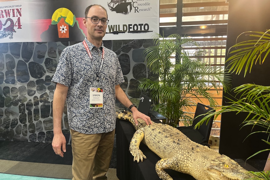 A man wearing a red lanyard and a bright patterned shirt stands next to a model of a crocodile.