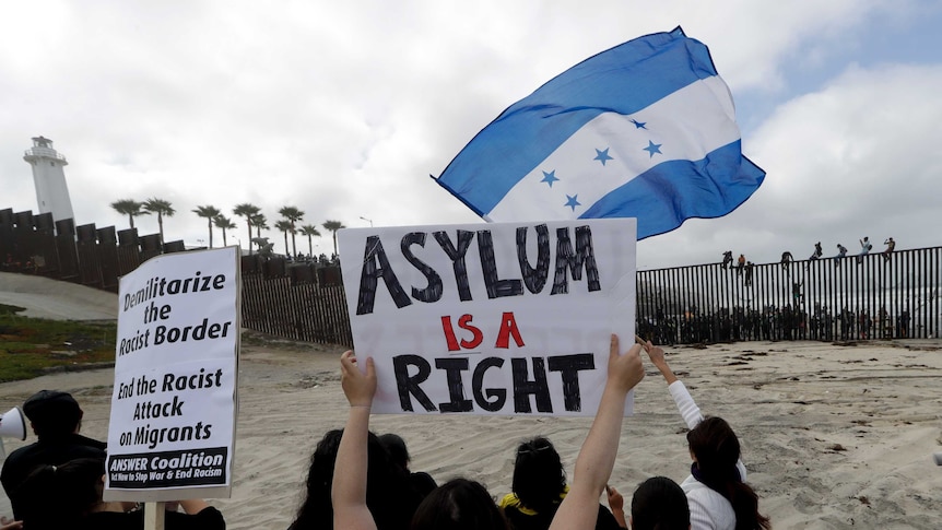 Migrants from both sides of the border rally. Some are sitting on top of the border wall, others have flags and placards.