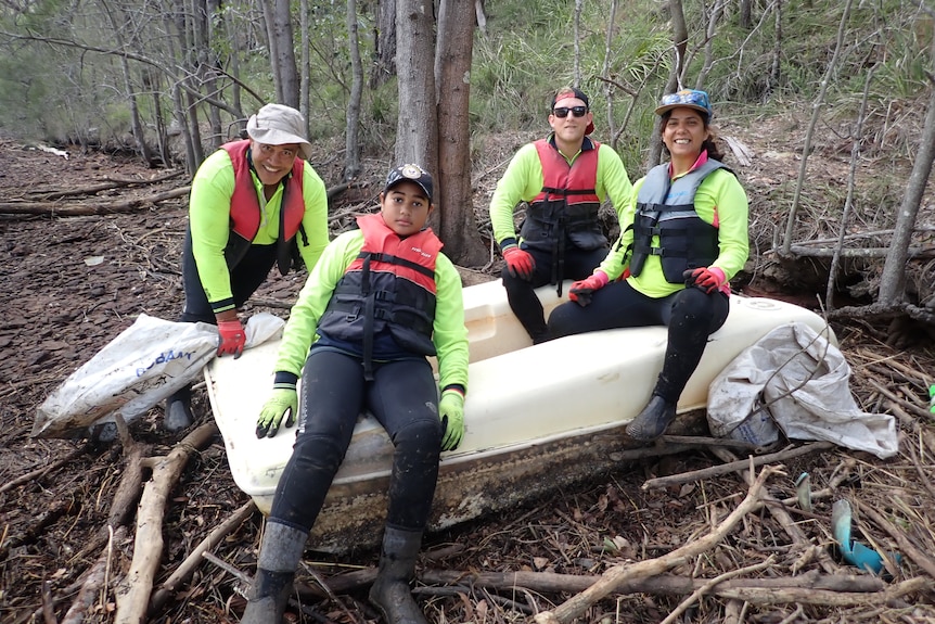 Four people sitting on an overturned boat in bushland.