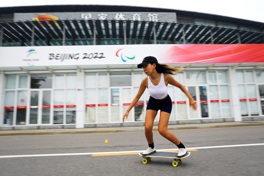 A woman riders a skateboard in front of a building that says Beijing 2022. 