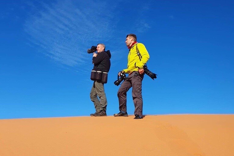 Two men stand on a sand dune with a blue sky behind them. 