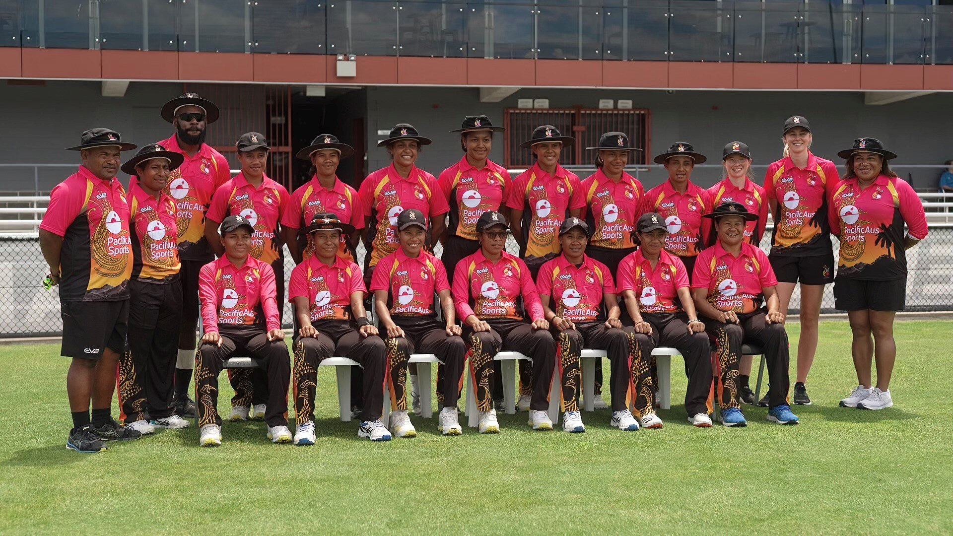 A female cricketer wearing red and black wide brimmed black hats sits on chairs for a formal team photo.