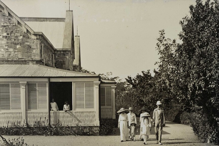 Black and white photo of a group of people walking away from an old brick house in early 1900s outfits.