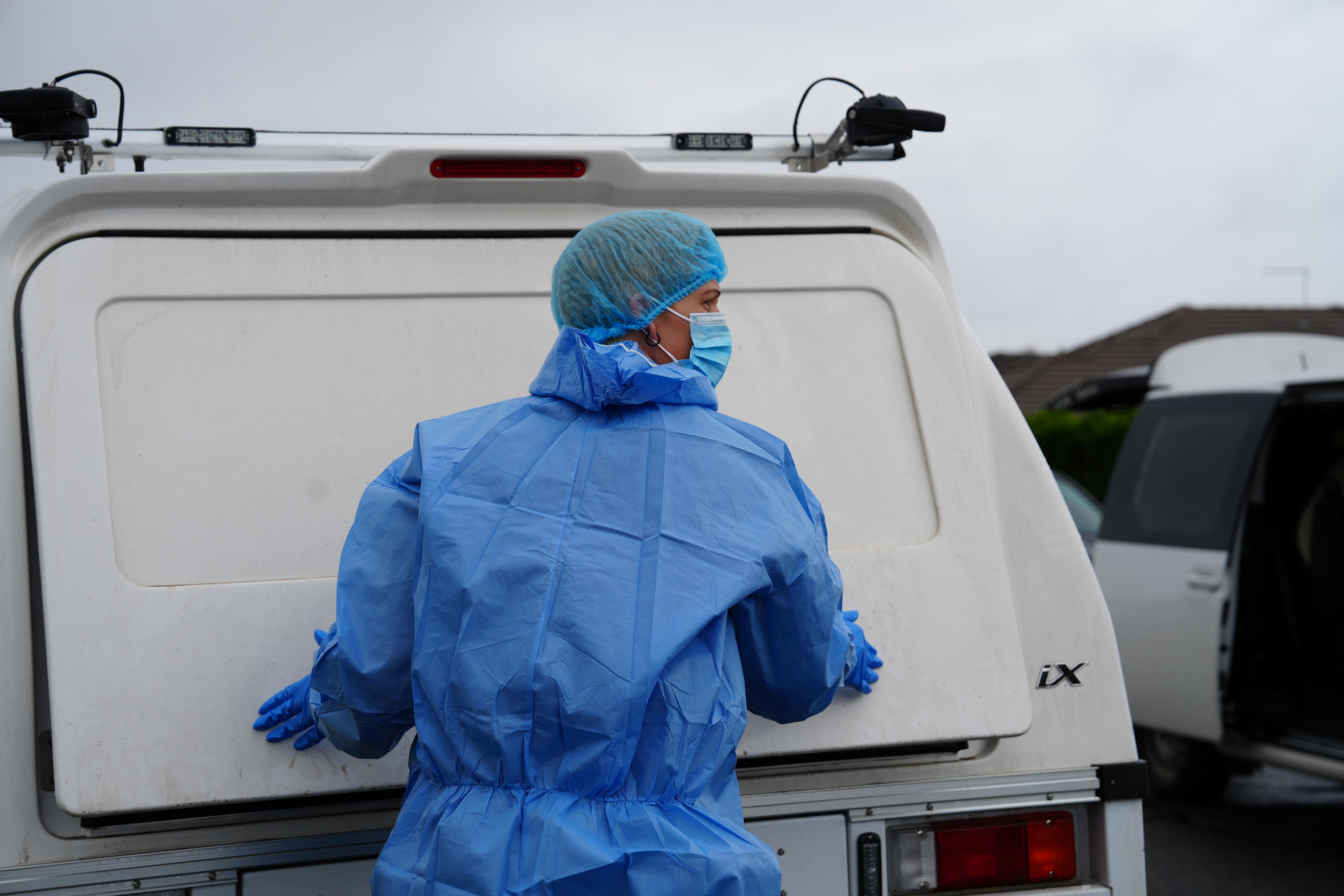 A person in blue protective scrubs, a mask and hair net stood behind a white utility.