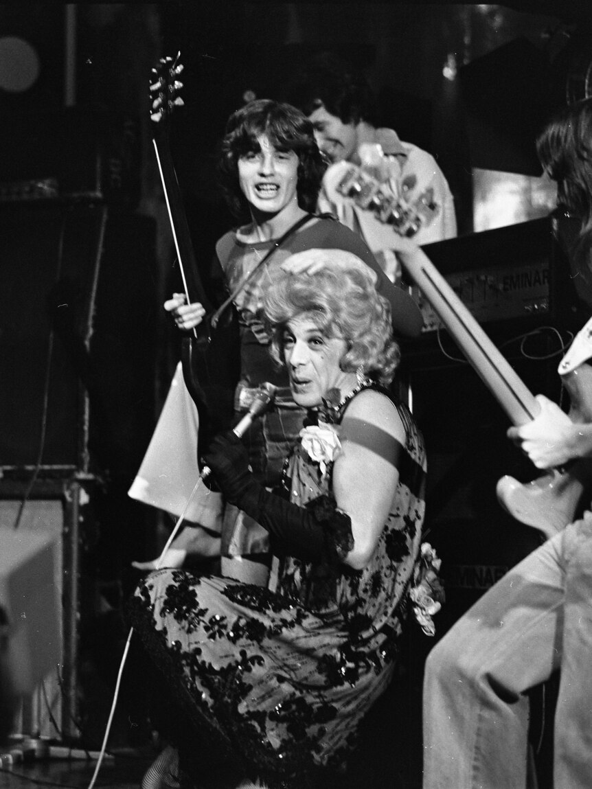 Black and white image Molly Meldrum with microphone in dress and women's wig next to AC/DC guitarists aughing