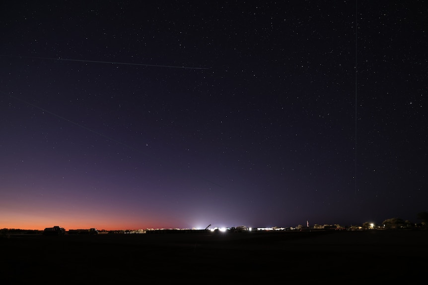 A twilight shot of stars and three satellites zooming across the skies with distant lights of the fly camp in the background