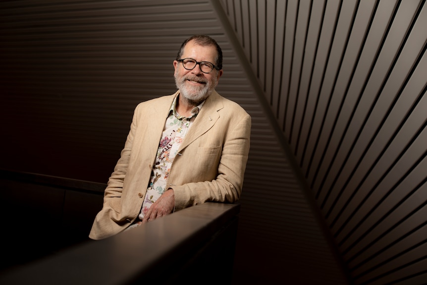 A man with a grey beard and glasses, wearing a beige suit and tropical print shirt, leans against a bannister