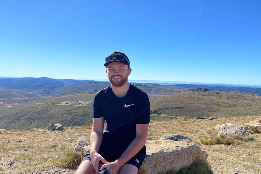 A man sits on a rock with rolling hills in the distance behind him.